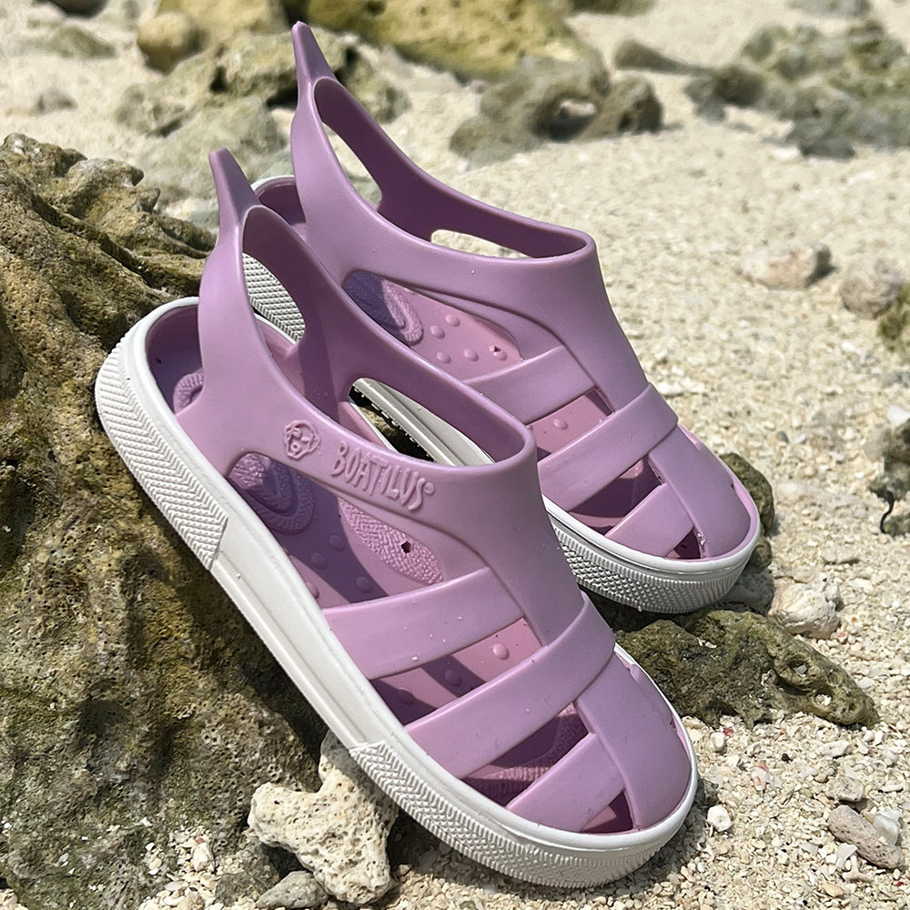 Image of a pair of Boatilus Bioty sandals in lilac resting on a rock on a sandy beach