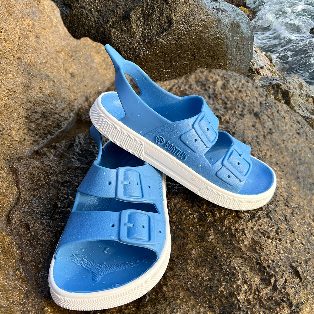 Image of a pair of Boatilus Irky sandals in jeans blue and white on a rock near the sea